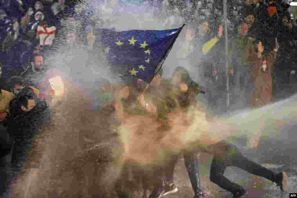 Ýewropa Bileleşiginiň baýdagyny göterýän demonstrantlara suw sepilýär. Protestçileriň we polisiýanyň arasynda çaknyşyklar 2023-nji ýylyň 7-nji martynda Tbiliside Gürjüstanyň mejlisiniň golaýynda boldy. Gürji polisiýasy paýtagt Tbiliside jedelli "daşary ýurt agentleri" kanunyna garşylyk bildirmek üçin köçä çykan müňlerçe adamlyk protestiň gatnaşyjylaryna garşy gözi ýaşardýan gazy we suw pürkýän gurallary ulandy. (AFP suraty)